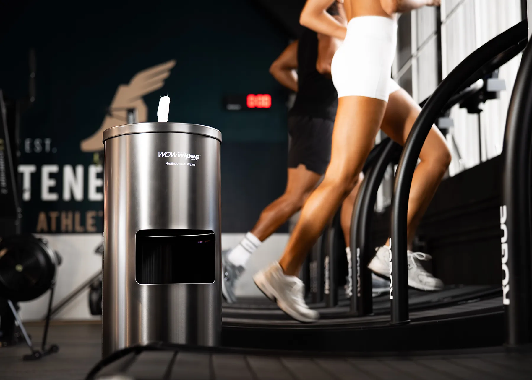 black wet wipes dispenser next to a man and women running in the gym.