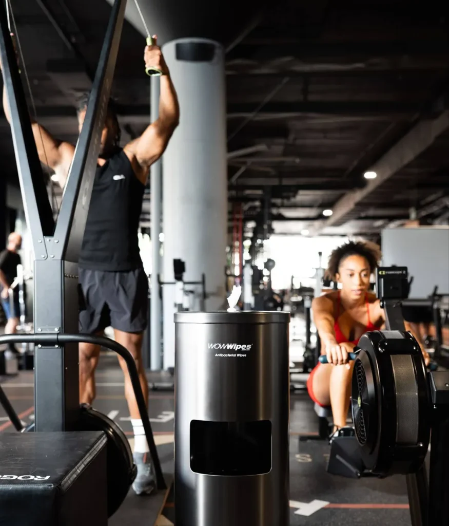 WOW Wipes® Cleaner for Gym Spaces: A Cleaner and Greener Facility Male and female using gym equipment with a black wet wipe black standing dispenser